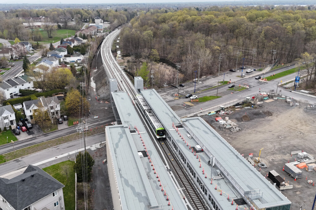 Vue aérienne d’une station de métro en construction, avec un train sur les rails, des routes, des maisons et une forêt arborée